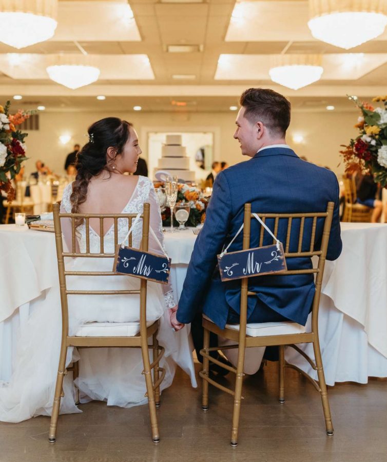 A bride and groom sitting at the sweetheart table, holding hands and smiling at one another.